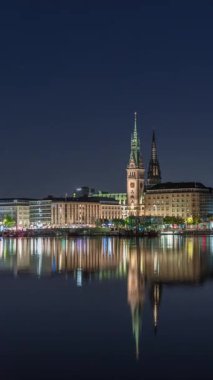 Binnenalster 'in Hamburg, Almanya' daki Belediye Binası ve Nikolai Kilisesi 'ndeki gece programı. Aydınlatılmış tarihi binalar yıldızlı bir gökyüzünün altındaki Alster Gölü sularına yansıyor. Manzaralı şehir manzarası