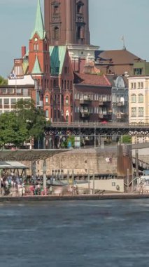 Hamburg limanına giriş ve çıkış yapan bir feribotun rıhtımdaki yolcuları ve merdivenleri gösteren zaman çizelgesi. Arka planda Elbe nehri boyunca tarihi ve modern binalar var. Almanya
