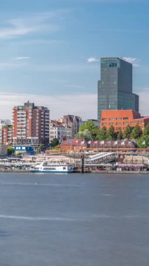 Hamburg limanına giriş ve çıkış yapan bir feribotun zaman çizelgesi. İskelede yolcuları var ve merdivenlerde ilerliyor. Arkaplanda Elbe nehri boyunca tarihi ve modern binalar var. Almanya