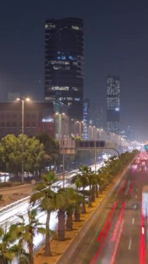 Suudi Arabistan, Riyad 'daki ikonik bükümlü gökdelenle King Fahd Yolu' ndaki yoğun karayolu trafiğinin hava takvimi görüntüsü. Aydınlanmış sokaklar ve ağaçlar. Dünyanın en uzun sekizinci büküm kulesi..