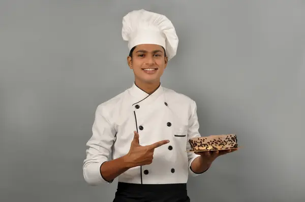 Portrait of a happy male chef dressed in uniform holding plate with Chocolate cake Pointing on Cake