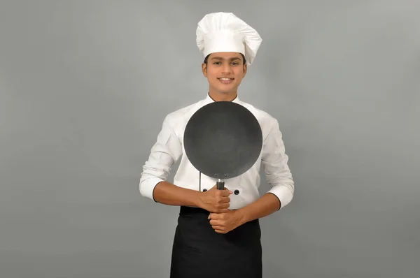 Confident smiling Indian chef smiling behind the frying pan, on grey background