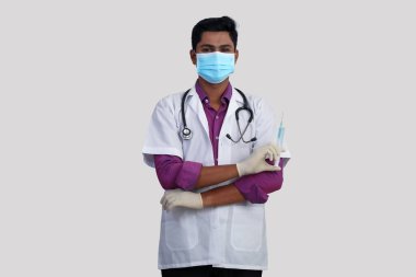 Yüz maskeli genç Hintli erkek doktor kollarını kavuşturmuş, tıbbi eldiven takmış beyaz arka planda izole edilmiş şırınga tutuyor. 