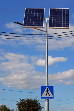 Arka planda güneş enerjisiyle çalışan sokak lambası ve yaya tabelası var.