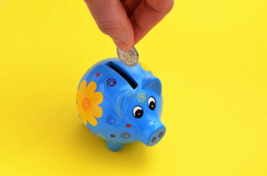 El, sarı arka planda mavi bir kumbara içine bozuk para indirir.