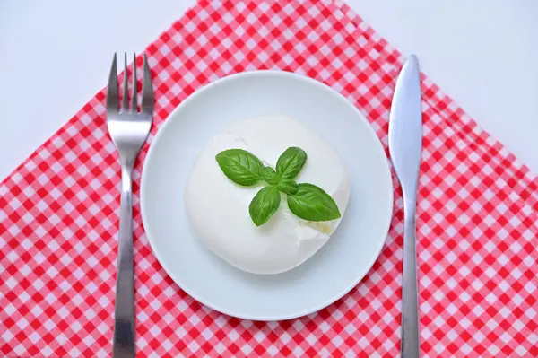 Kırmızı ve beyaz masa örtüsü üzerinde çatal ve bıçakla servis edilen fesleğenli burrata peyniri.