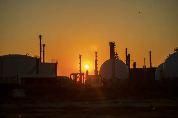 silhouettes of structures, pipes, oil storage tanks, pipelines of a petrochemical plant in the backlight of the sun at sunset and a sun disk