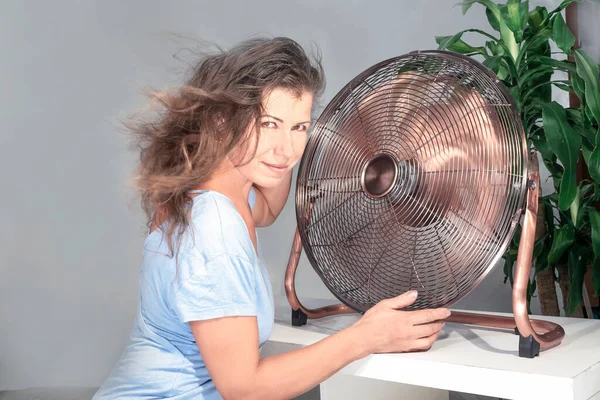 woman holding air cooling fan smiles enjoying the wind from the blades in the apartment. Cooling the air in the summer heat.