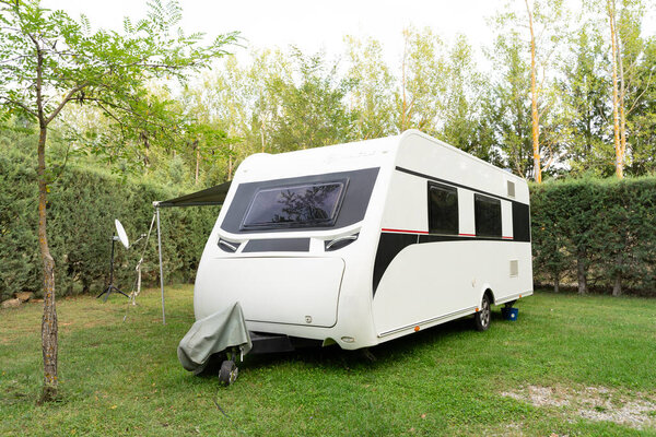 A white trailer,a motorhome for travel,a motorhome,camper,trailer without a car against a background of beautiful bushes stands on short green lawn grass.Technologies of transport, means of travel.