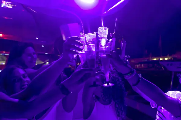 girls with raised hands with glasses with champagne alcohol connect glasses,laugh and pose for the camera in the light of neon blue lights illuminating the territory of the catamaran yacht. Yaht party.