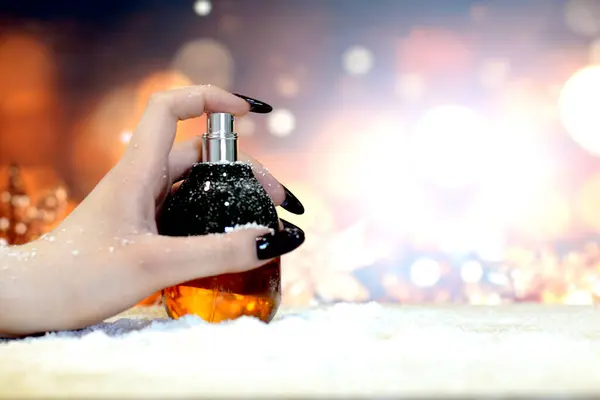 A woman\'s hand with gel long black nails holds a bottle of perfume on a festive background of blurred lights in shades of gold,elegant brown with sparkle. Presentation of perfumes and women\'s manicure.