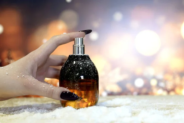 A woman's hand with gel long black nails holds a bottle of perfume on a festive background of blurred lights in shades of gold,elegant brown with sparkle. Presentation of perfumes and women's manicure.