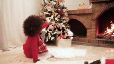 Koyu tenli küçük bir kız süslü bir Noel ağacına hayran ve şöminenin yanındaki oyuncağı işaret ediyor. Evde Noel zamanı. Noel 'i kutlamak.