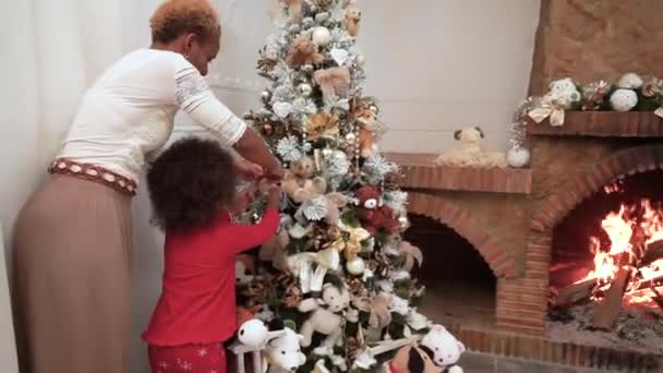 アフリカの暗い肌のママと子供は火の暖炉の隣に飾られたクリスマスツリーにクリスマスボールを掛け 楽しんでいます クリスマスツリーを飾る家族 クリスマスタイム — ストック動画