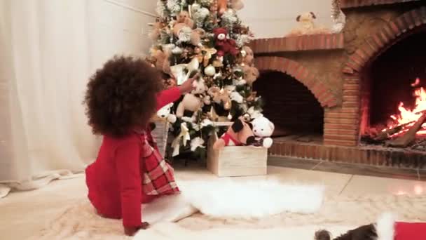 一个黑皮肤的小女孩欣赏一棵装饰过的圣诞树 并指了指壁炉边的一个有火的玩具 圣诞节在家里 庆祝圣诞节 — 图库视频影像