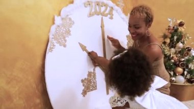 Afrikalı anne ve küçük kız büyük bir dekoratif saatin ellerini şeftali rengi bir Noel ağacının arka planında 2024 numaraya doğru hareket ettiriyor.