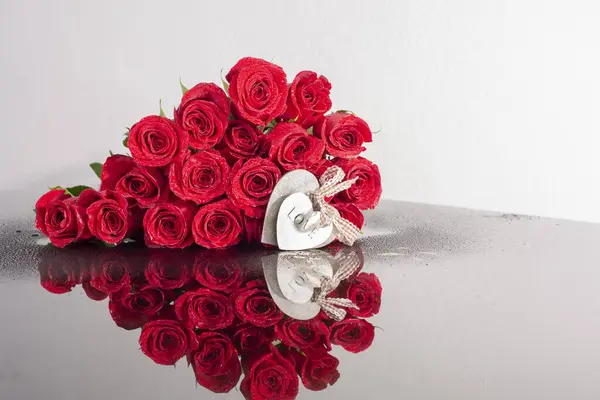 Floral heart of roses with water drops on a mirror table with a beautiful reflection and decorative hearts. Symbol of love and Valentine\'s Day. Red roses are collected in the shape of a heart.