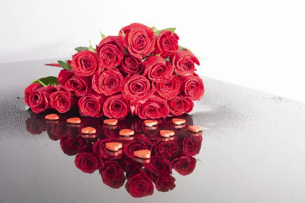 Floral heart of roses with water drops on a mirror table with a beautiful reflection and decorative hearts. Symbol of love and Valentine\'s Day. Red roses are collected in the shape of a heart.