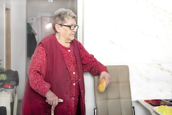 A sweet grandmother with a stick waits for breakfast with a loaf of bread in her hand at a nursing home. Nutrition for the elderly, diet in old age, getting help.