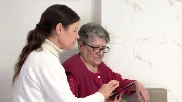 中年の女性は 携帯電話の設定を高齢のおばあさんに説明します 古い人々によって新しい技術を学ぶ 若者の助けを借りて携帯電話を研究している老人 — ストック動画