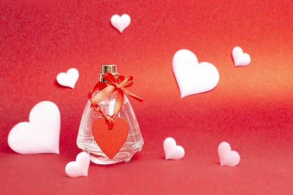 A bottle of perfume with a red festive ribbon with a heart on a red background with hearts. Gift card for Valentine\'s day. Cosmetics as a gift for mother\'s day, birthday. PHOTO CREATED USING A CAMERA