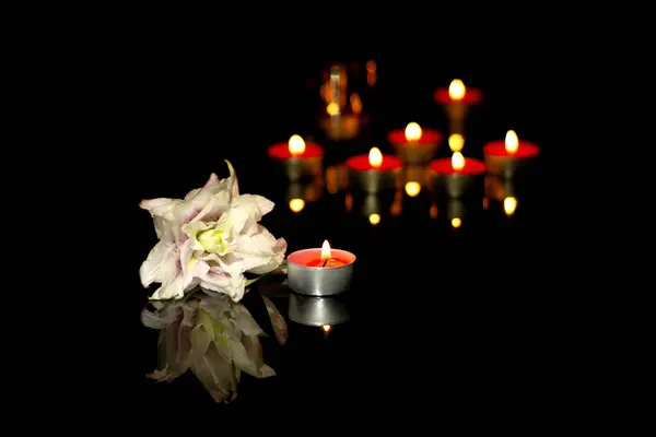 White lily and burning candles In the silence of the darkness of a funeral on a black plaque with the reflection of lights. Sad mourning background