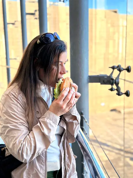 A hungry woman eats a hamburger sandwich near the window outdoors. Takeaway, fast food. Takeout, street food