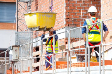 Koruyucu, güvenlik ekipmanı kullanan bir işçi inşaat halindeki bir binanın yüksekliğinde vinç zincirinde beton bir karışım bulunan bir konteynerin kaldırılmasını yönetir. Fotoğraf, bir fotograf NIKON 610 kullanarak yaratıldı.
