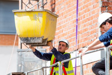 Koruma ve güvenlik ekipmanlarında çalışan bir işçi, inşaat halindeki bir binanın yüksekliğinde vinç zincirinde beton bir karışım bulunan bir konteynerin kaldırılmasını yönetir. Fotoğraf, bir fotograf NIKON 610 kullanarak yaratıldı.