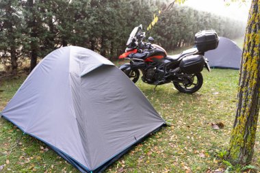 Park etmiş bir motosikletle yaz ormanının gölgesinde çadır kurmak. Eğlence için ekipman ve aksesuarlar. Orman Çevre dostu Geçici Kumaş Mobil Evi