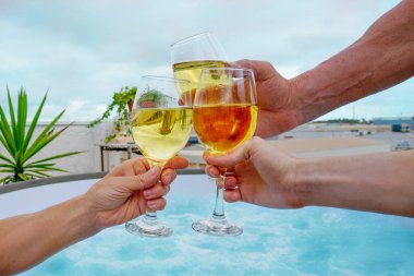 Üç kişinin ellerinin şarap bardaklarını tutarken, kadeh tokuştururken, gök mavisi suyun arka planına köpüklü yüzme havuzu jakuzisiyle vururken, terasta.