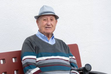 Şapkalı yaşlı adam bankta oturuyor, beyaz arka planda bastonun yanında gülümsüyor. Emekli büyükbaba dinleniyor, mutlu yaşlılar.
