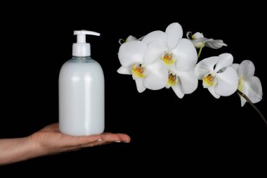 Siyah arka planda beyaz orkideli bir kadın eli üzerinde sahte şeker şişesi. Cilt bakımı için kozmetik ürünleri sunumu. El kremi ve hijyen ürünleri. Şefkat ve saflık