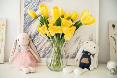 İki pelüş tavşancık sarı laleleri ve nergisleri olan bir vazonun yanında, arka planda hafif dekorlu bir rafta oturuyor. Paskalya için yapay çiçekli ve tavşanlı ev dekoru.