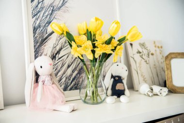 İki pelüş tavşancık sarı laleleri ve nergisleri olan bir vazonun yanında, arka planda hafif dekorlu bir rafta oturuyor. Paskalya için yapay çiçekli ve tavşanlı ev dekoru.