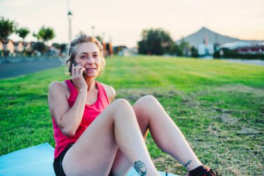 Pembe halterli ve mavi hasırlı bir kadın çimlerin üzerinde oturuyor. Cep telefonuyla konuşuyor.