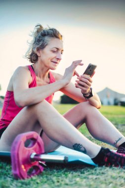 Pembe dambılı ve mavi minderli çimenlerde oturan bir kadın. Cep telefonunu kullanıyor.