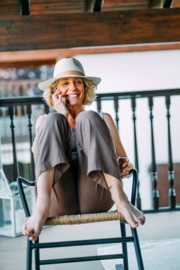 Bir kadın sandalyede oturuyor ve cep telefonuyla konuşuyor. Şapka takıyor ve yüzünde bir gülümseme var.