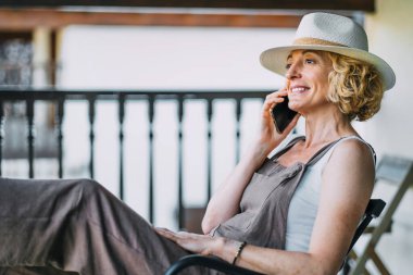 Bir kadın sandalyede oturuyor ve cep telefonuyla konuşuyor. Hasır bir şapka ve beyaz bir gömlek giyiyor.