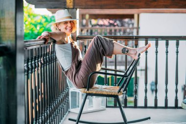 Bir kadın balkonda bir sandalyede oturuyor, hasır şapka ve beyaz gömlek giyiyor. Gülümsüyor ve manzaranın tadını çıkarıyor.