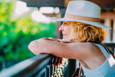 Hasır şapka takan bir kadın gülümsüyor ve korkuluklara yaslanıyor. Elinde bir çanta var ve manzaranın tadını çıkarıyor.