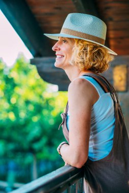 Hasır şapka takan bir kadın gülümsüyor ve korkuluklara yaslanıyor. Elinde bir çanta var ve manzaranın tadını çıkarıyor.