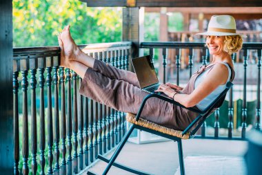 Bir kadın balkonda sallanan bir sandalyede oturuyor, hasır şapka takıyor ve dizüstü bilgisayar kullanıyor. Rahatlama ve rahatlama kavramı, kadın laptopuyla çalışırken dışarıda vakit geçirmeyi seviyor.