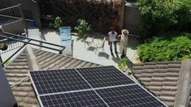 Havadan bakıldığında çatıda güneş panelleri ve güneşli bir arka bahçede iki kadın görülüyor. Biri sandalyede dinleniyor, diğeri de çiçekleri suluyor. 4 bin. Görüntü çevre dostu çevreyi vurguluyor..