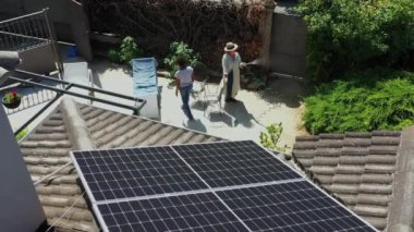 Havadan bakıldığında çatıda güneş panelleri ve güneşli bir arka bahçede iki kadın görülüyor. Biri sandalyede dinleniyor, diğeri de çiçekleri suluyor. 4 bin. Görüntü çevre dostu çevreyi vurguluyor..