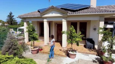 Bir kadın, çatıda güneş panelleri olan güneşli bir bahçede bitkileri suluyor. 4 bin. Sahne, çevre dostu bir ortamda geçen bir bakım ve sürdürülebilirlik anını yakalıyor..