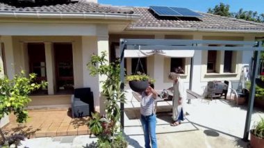 Güneşli bir bahçede bitkilerle ilgilenen iki kadın var. 4 bin. Sahne, çatıda güneş panelleri olan ve saksı bitkileriyle çevrili bir evin önünde geçiyor..