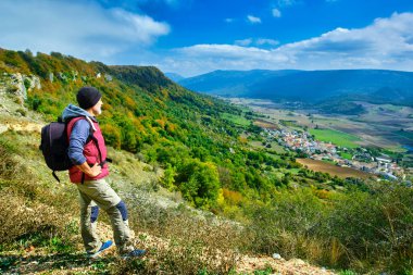 Bir yürüyüşçü, sonbahar mevsiminde göz kamaştırıcı bir vadiye bakan, canlı yeşil, sarı ve turuncu yapraklı bir dağ yolunda duruyor. Urbasa-Andia Doğal Parkı ve Eulate Köyü. Navarre, İspanya.