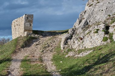 Kayalık arazi ve yeşillik parçalarıyla çevrili eski bir taş yapıya doğru uzanan toprak bir yolun görüntüsü. Vitoriano, Araba, Bask Ülkesi, İspanya.