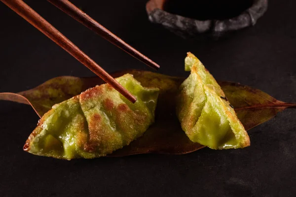Green vegetable gyozas picked up with chopsticks Japanese steamed, delicious Japanese food with black background and decorated with leaves and a wicker basket, for oriental and Asian food restaurant.